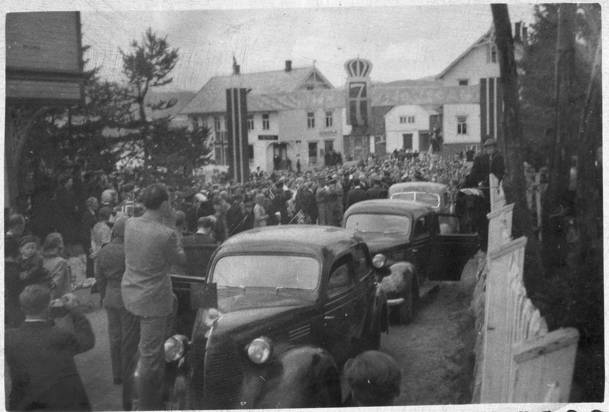 Folk og biler på gata i Sjøvegan under kongebesøket i 1946. Flagg og H7-monogram i bakgrunnen.