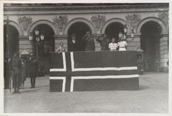 Kong Haakon stående på scene dekorert med det norske flagget