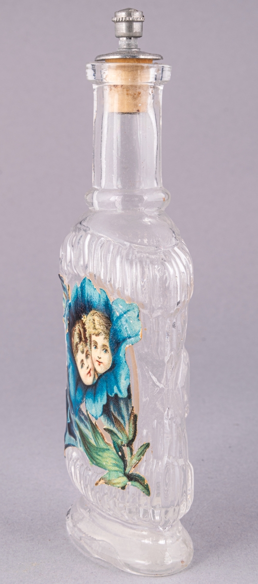 Parfymflaska, med glasknopp och gult band runt knoppen. Etikett med blomma och två ansikten.