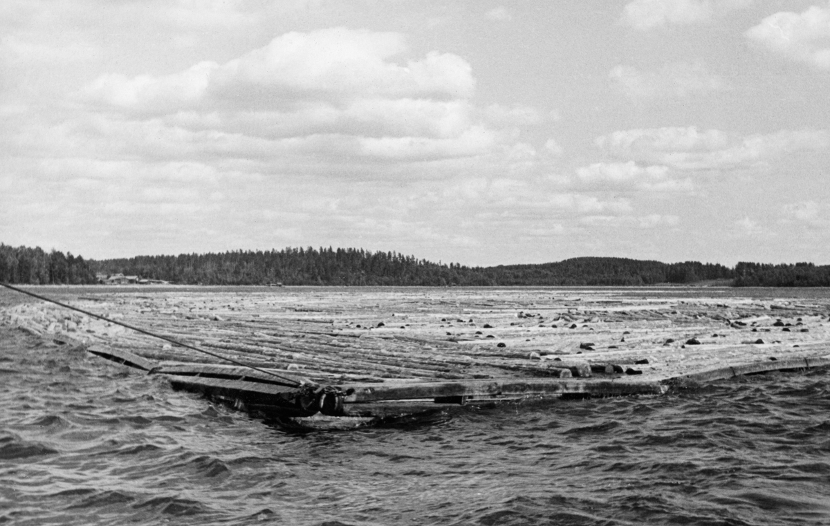 Rringbom under buksering på den finske innsjøen Kirkkojärvi, antakelig bak slepebåten «Leini». Ringbommen er et flytende stengsel, laghd av sammenlenkete doble bjelker og med et solid jernbeslag som slepetrossa er festet i. Vinkelen mellom vannspeilet og trossa tyder på at avstanden mellom fartøyet og trossa var forholdsvis kort. I Norge var man opptatt av at det skulle være en viss avstand mellom de to enhetene, slik at man unngikk at «propellvannet» presset bommen bakover samtidig som slepebåten forsøkte å trekke forover. Stedsnavnet Kirkkojärvi er det flere av de finske sjøene som har, og vi vet ikke hvilken av dem dette dreier seg om. Fotografiet ble tatt under en ekskursjon for norske fløtingsfunksjonærer i Sør-Finland i 1934.