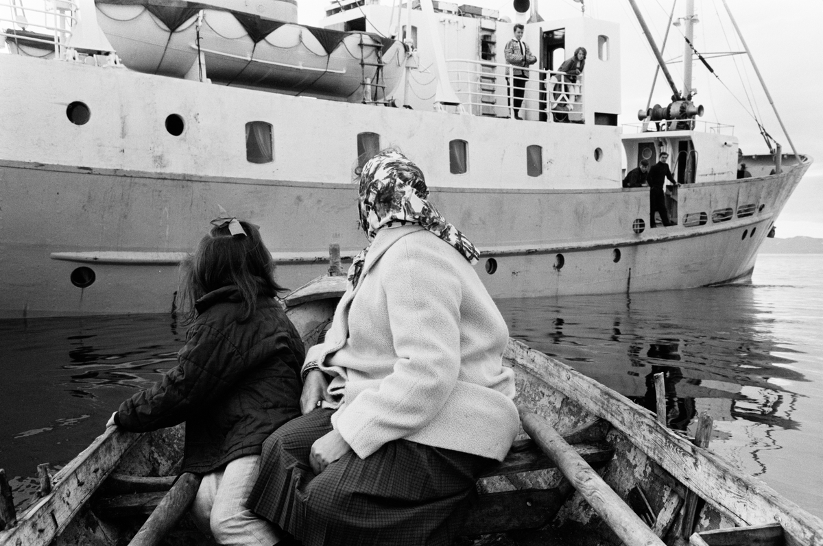 Jente og eldre kvinne i liten båt ved siden av en stor båt.
