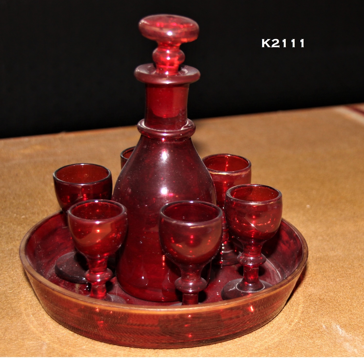 K2ll1, a—h. 
Likørkaraffel med 6 glass på brett, 1850—årene. Rubinglass, bøhmisk. 
a) Brett, sirkulært med skrå karm og forgylt kant.
b) Karaffel.
Runde skuldre, en ring om den smale hals, munningsbrem og propp. 
e-h) Glass, 6 stk. Rund fot, balusterstett, klokkeformet cupa.
