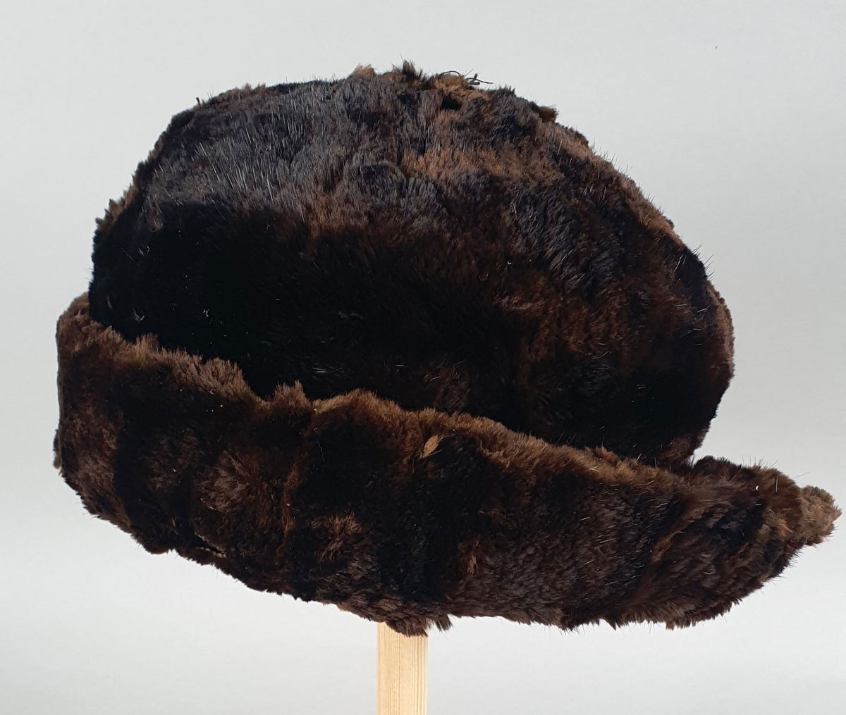 Mørkebrun hatt av pels (mink?), fôret med mørkebrun bomull. Brem som brettes opp langs pullen, med en splitt midt bak. Skinnet er revnet på flere steder.