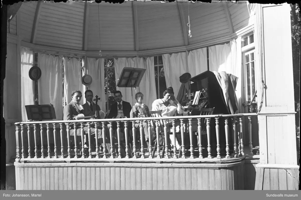 Musiker sitter med sina instrument i en paviljong, tre män, en kvinna och en ung pojke. Kvinnan finns även med på bild SuM-foto000547.
