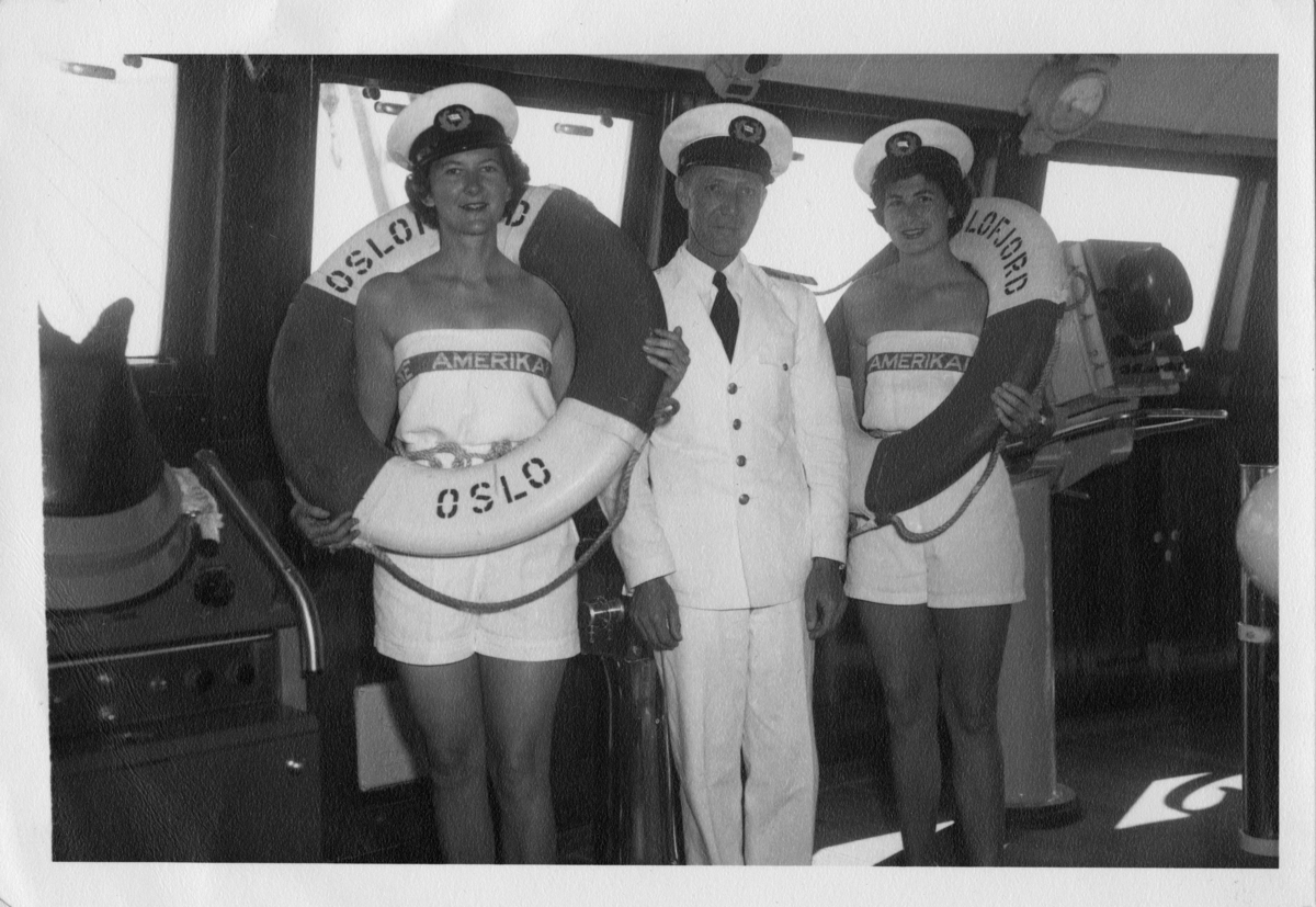 Merket "Passengers". To kvinner med kapteinslue og redningsbøye fra NAL-skipet OSLOFJORD, på broen sammen med kapteinen.