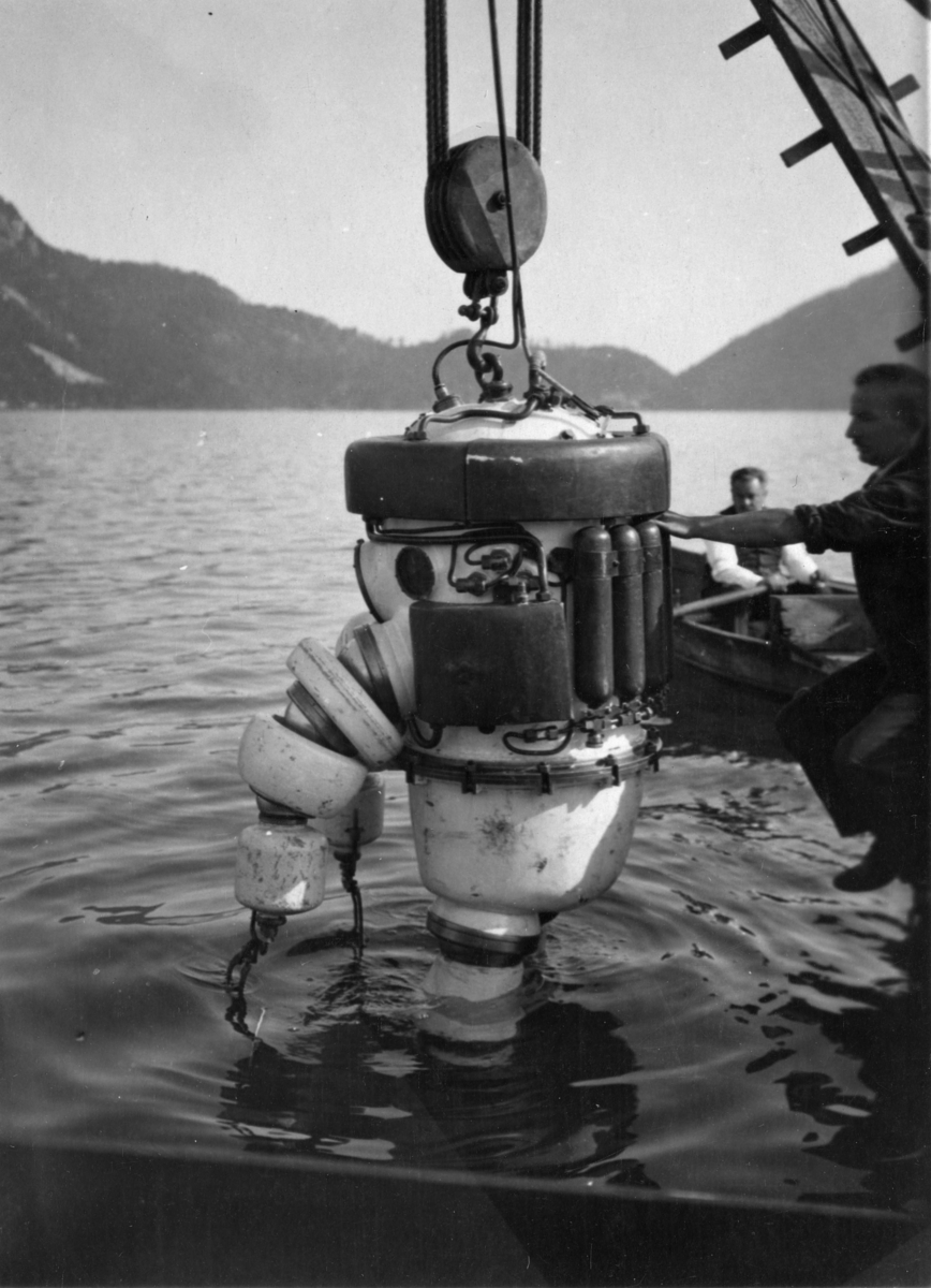 Dykker i panserdrakt senkes ned i vannet med vinsj, mann i robåt.