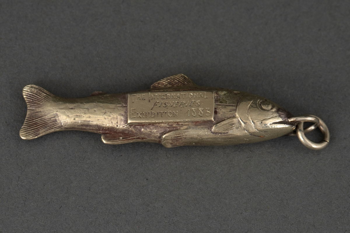 Fiskeformet merke for jurymedlem ved The International Fisheries Exhibition 1883. Hvit metall, antagelig forsølvet. Ring for feste til kjede eller lignende.
