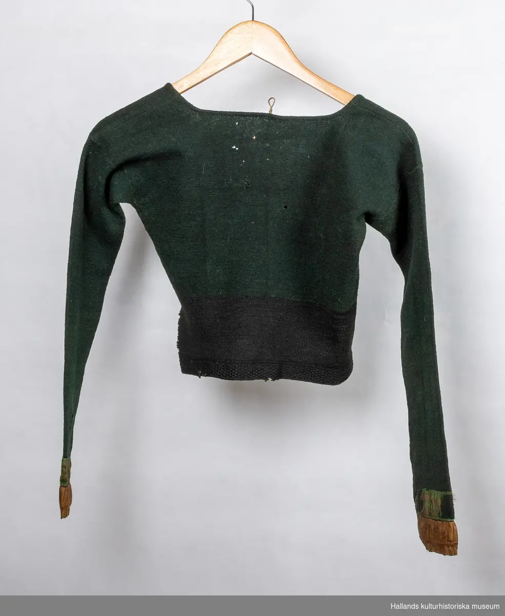 Grön stickad tröja i ull med svart nederkant. Sidenband på ärmlinningarna. Längd från axeln 40 cm. Längd på ärmen 50 cm. Midjevidd 66 cm.