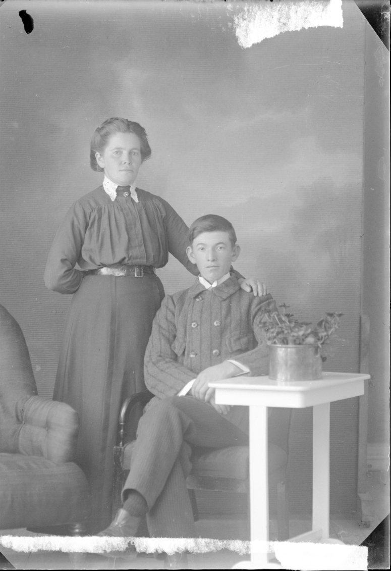 En ung man sitter vid ett bord. En ung kvinna står intill, håller sin vänstra hand på pojkens axel.