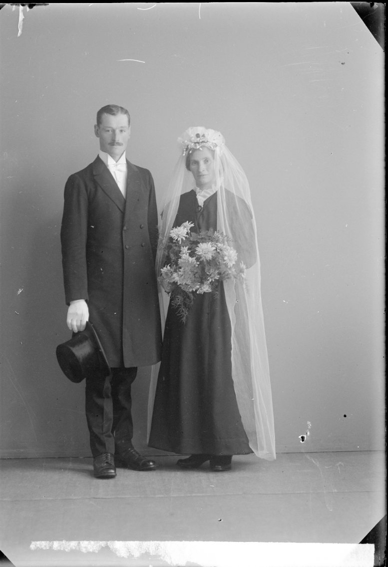 Ett brudpar. Bruden i svart. Brudgummen håller den höga hatten i höger hand.