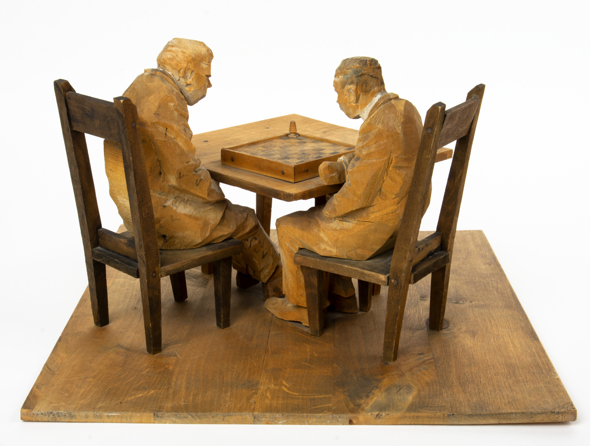 Skulptur av trä av föreställande två schackspelare sittande på var sin stol vid ett bord.