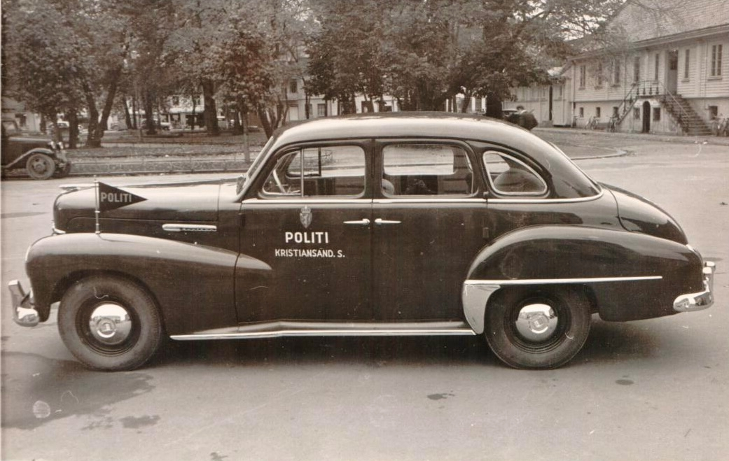 En personbil K-7, Opel Kaptein 1954-modell merket "Politi Kristiandsand S." med registreringsnummer K-7 og politiskilt formet som en vimpel festet over venstre forhjul. Bilen står parkert foran en bygning. Bilen er fotografert både forfra og fra siden.