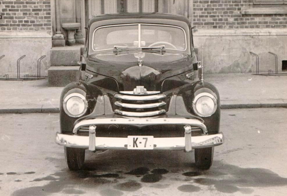 En personbil K-7, Opel Kaptein 1954-modell merket "Politi Kristiandsand S." med registreringsnummer K-7 og politiskilt formet som en vimpel festet over venstre forhjul. Bilen står parkert foran en bygning. Bilen er fotografert både forfra og fra siden.