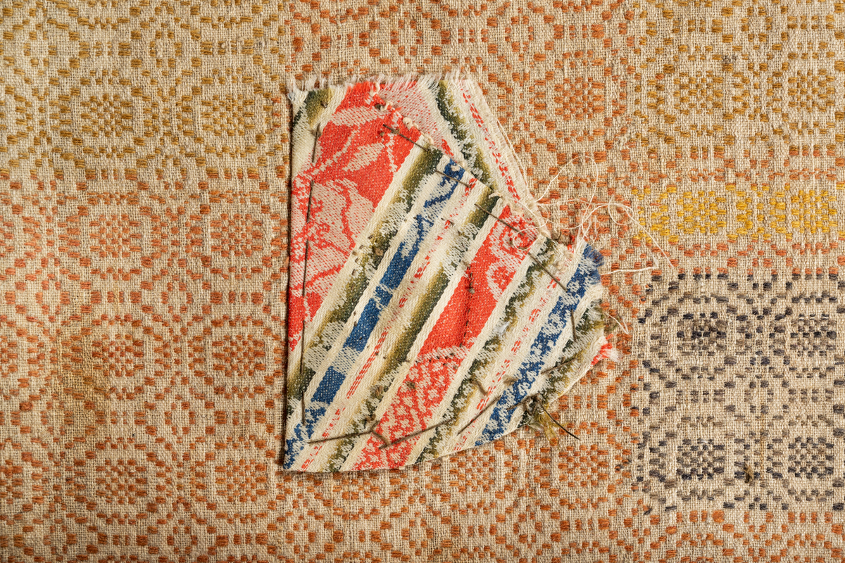 Täcke med varp av oblekt lin med inslag i brunt (gråblått enligt  katalogkorten), beige, gulrött och gult ullgarn. Två våder ihopsydda.