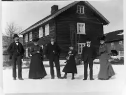 Seks personer avbildet foran et tømret våningshus i 2,5 etas