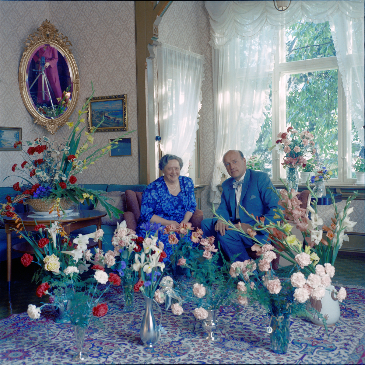 Eilif og Lilly Schrøder omgitt av blomster i anledning deres sølvbryllup. Fotografen, Rakel Schrøder, vises delvis i speilet.