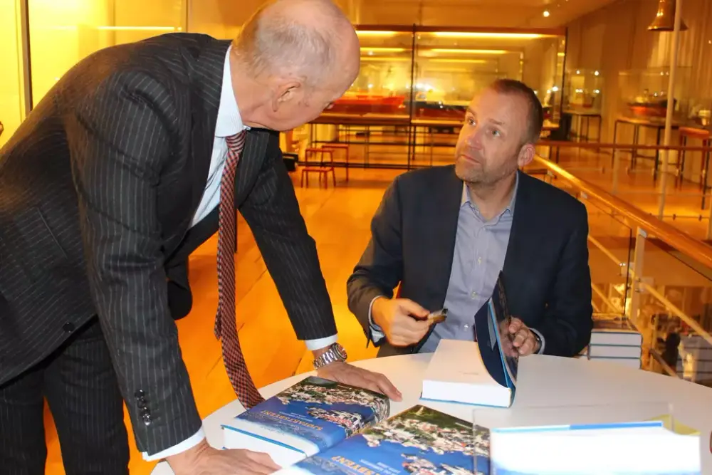 Forfatter Stig Tenold signerer bøker og prater med en gjest som skal kjøpe boken. I bakgrunnen ses museets utstilling. På bordet ligger bøker i stabler.