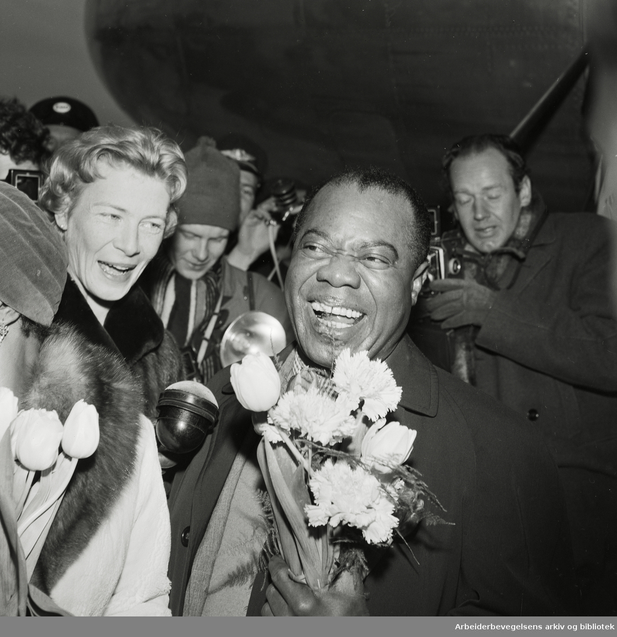 Louis Armstrong ankommer Oslo via Fornebu flyplass 2 februar 1959. Fra venstre: Lucille Wilson (hans kone), nrk-reporter Unni Børde og Louis Armstrong..