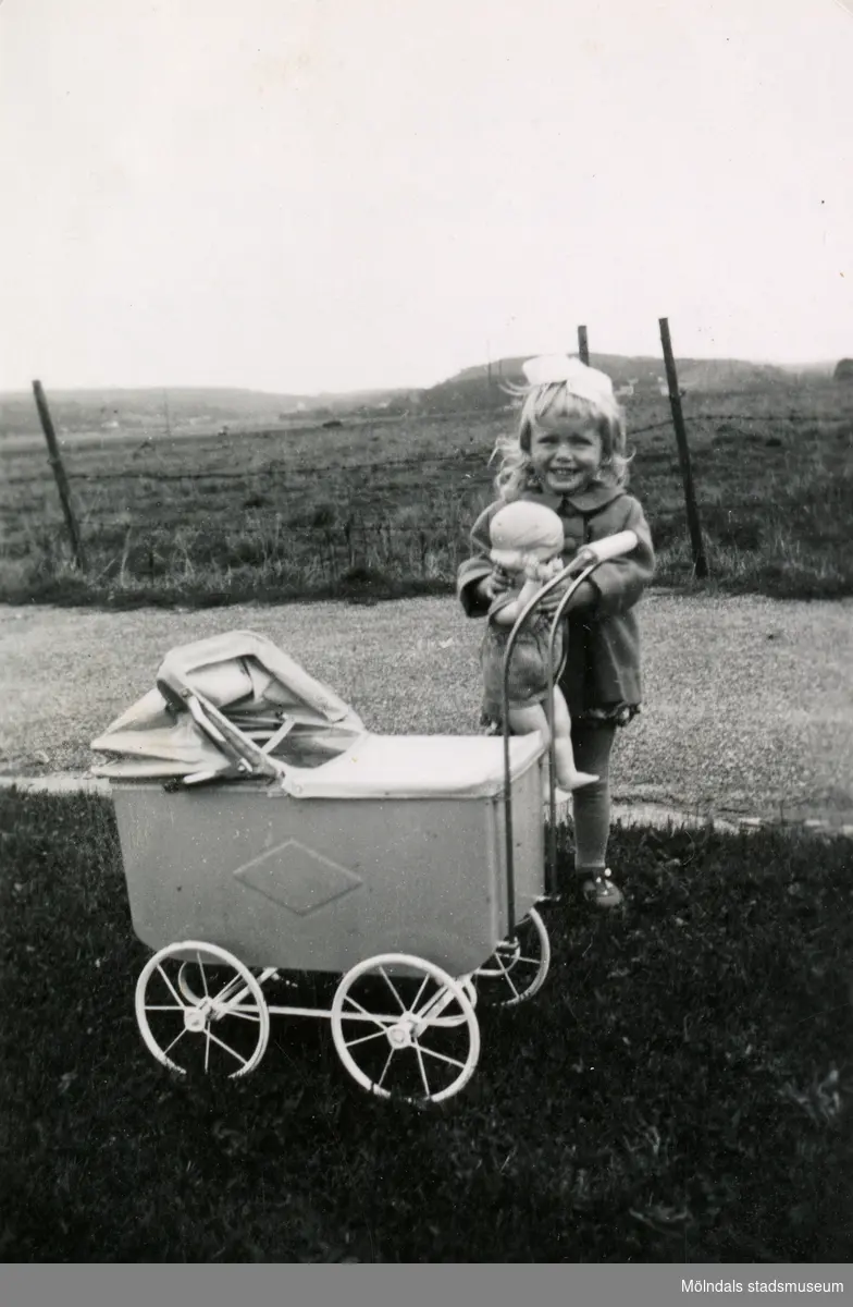 En liten flicka står med sin docka och dockvagn. Namn, årtal och plats är okänt.