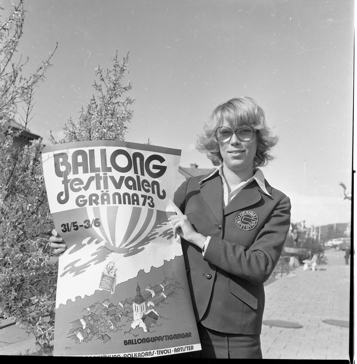 En ung kvinna med glasögon håller upp en affisch om "Ballongfestivalen i Gränna 73".