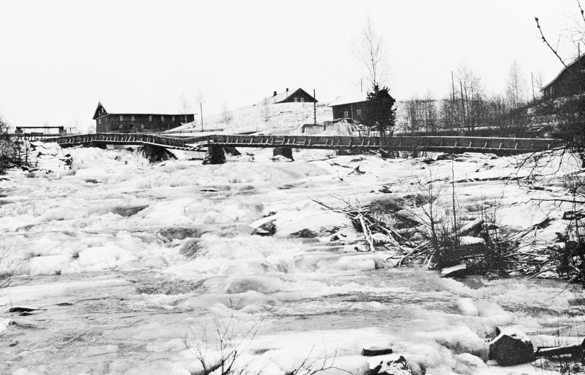 Fra Kråkfossen eller Krokfossen i elva Leira, som renner gjennom Nannestad kommune på Romerike. Fotografiet er tatt i motstrøms retning en vinterdag i 1939, da til og med vannet i fossen stort sett var frosset til is. Vi ser ei renne som skrår over fossen, på murte fundamenter som gav forholdsvis god klaring til underliggende is og vann. Et av spennene var imidlertid knekt. I bakgrunnen til venstre ser vi noe som kan ha vært brua på en reguleringsdam, til høyre ser vi driftsbygningen og våningshuset på en gard.