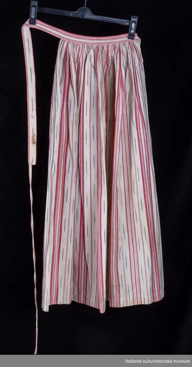 Randigt midjeförkläde i bomull. Vävt i vitt med ränder i rött (rosa) och ikatränder i vitt och blått. Påsydd linning i samma tyg med smalt band i vitt och rött. 