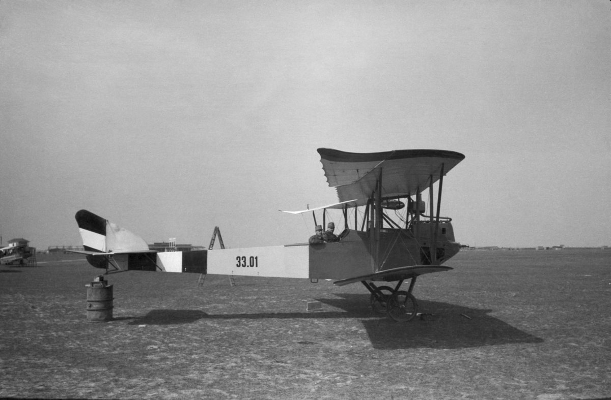 Militärt flygplan Aviatik B.III märkt 33.01 tillhörande Österrike-Ungern står på ett flygfält. Två män sitter i planet. Vy från sidan.