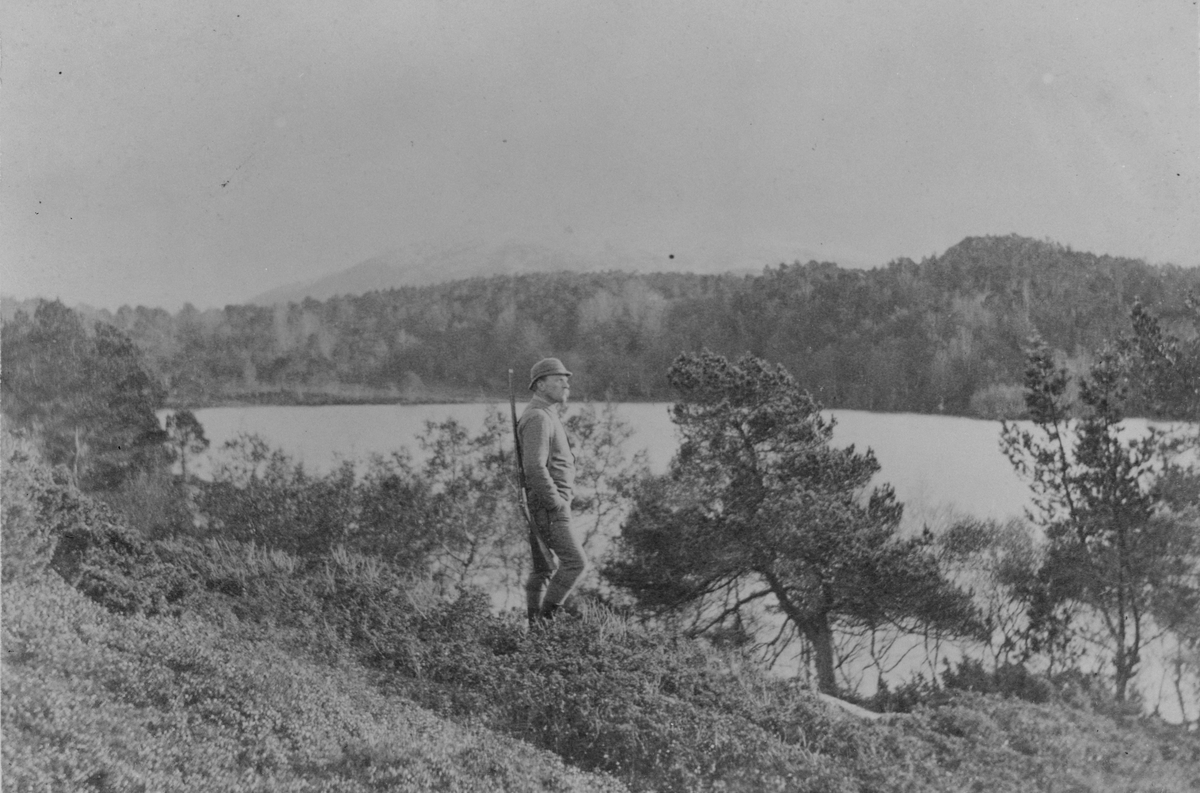 Olaf Løchen utendørs med jaktklær og gevær. Håvikfjellet i bakgrunnen, Kløfthaugen til høyre, Aunvann til venstre.