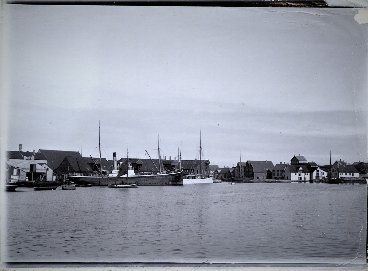 Et dampskip, en seilbåt og og diverse mindre båter ved Kortanes, Risøy i Haugesund. Til venstre ses Zakariassens bødkerverksted. To hus mot høyre står det H. M. Wrangell lager. 