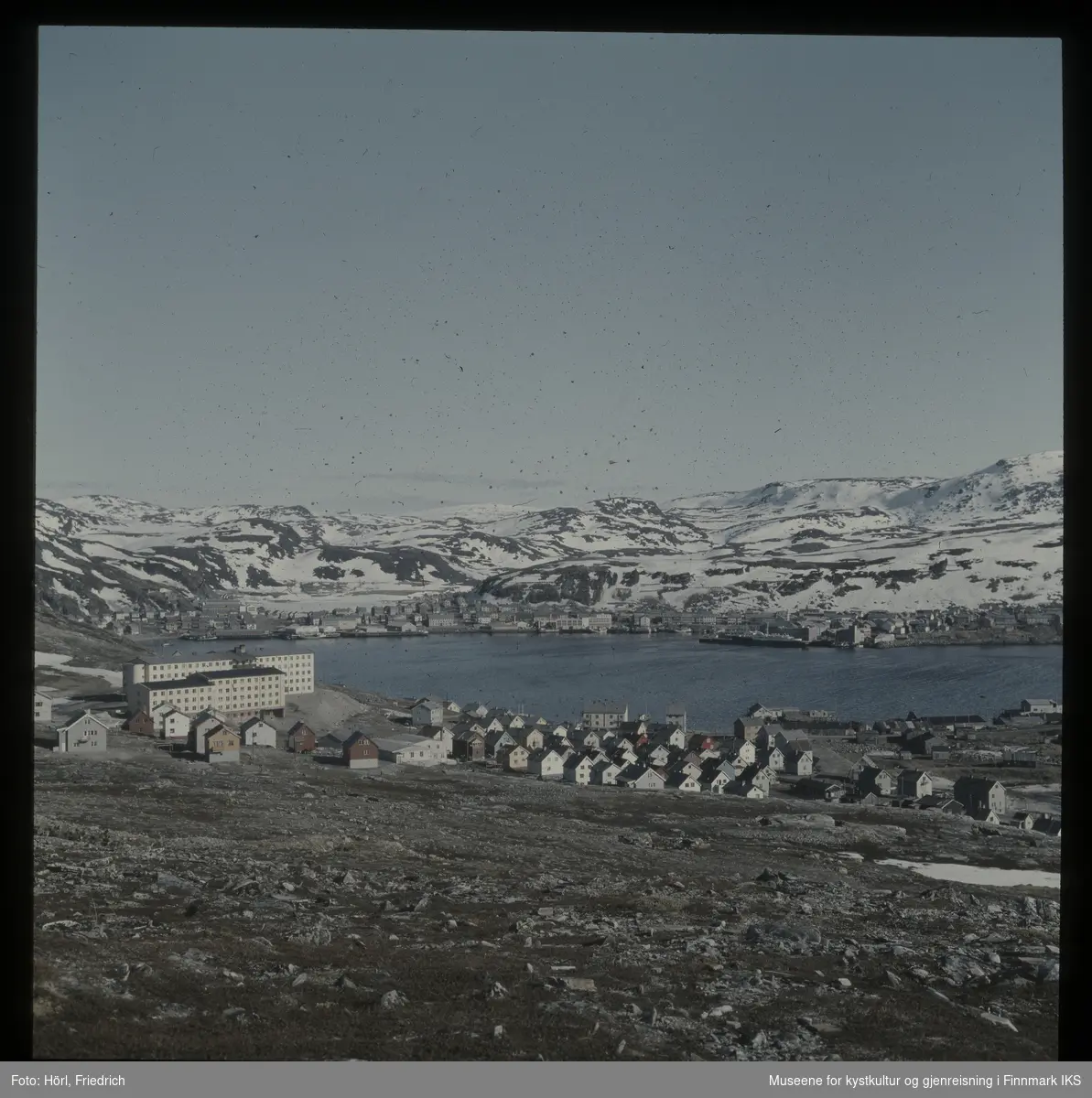 Utsikt over Hammerfest med sykehus og annet gjenreisningsbebyggelse i forgrunn. Bildet kan være tatt i våren 1958. Midt i bildet ser man havnebukta i Hammerfest, Mollaområde og byens sentrum bak den. På Storvannet og fjellene ligger det fortsatt snø.
