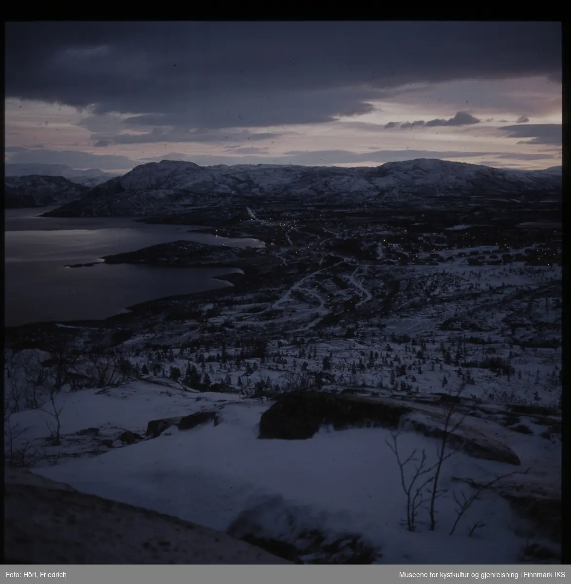 Utsikt fra et fjell på fjord og omliggende fjell. Landskapet er fortsatt dekket med snø og ligger i grålysningen. Området som vises er mest sannsynlig Bossekop ved Alta i Finnmark i vinteren 1972/1973.