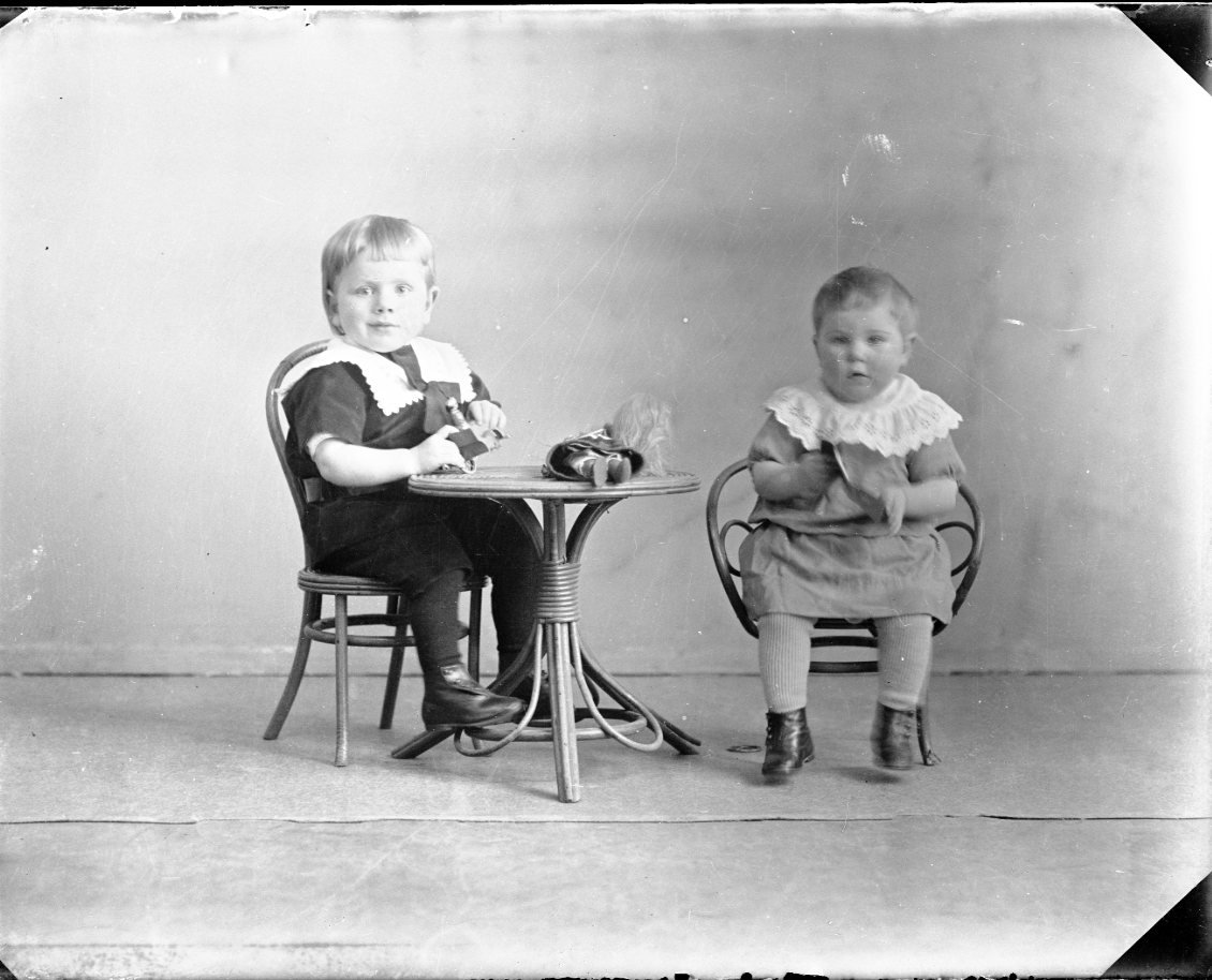 Två små syskon sitter vid en barnmöbel med leksaker, bland annat en docka.