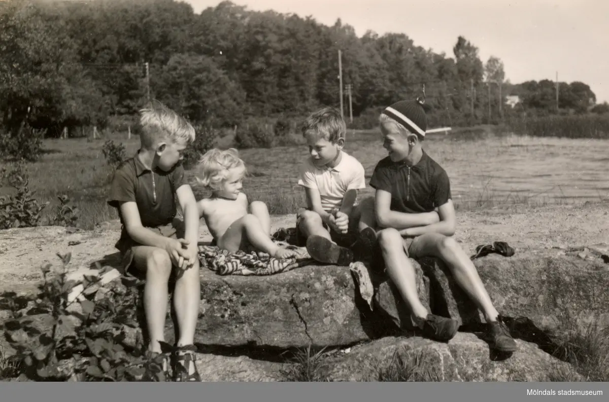 Eva Pettersson (född 1944, gift Kempe) sitter på en sten tillsammans med tre äldre pojkar (lekkamrater) vid Stenbryggan, Stretereds badplats, Tulebosjön år 1946.