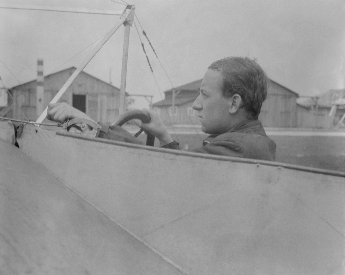 Edmund Sparmann sitter i förarsits på ett flygplan i Österrike-Ungern. Byggnader i bakgrunden. Vy från sidan.