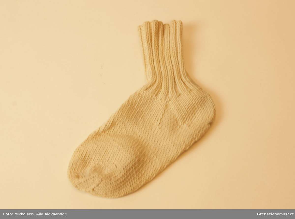 Gjenstanden er en enslig hvit sokk av håndstrikket bomull. Vrangbord to rette, to vrange. Stoffet er litt stivt, men ser relativt ubrukt ut.
