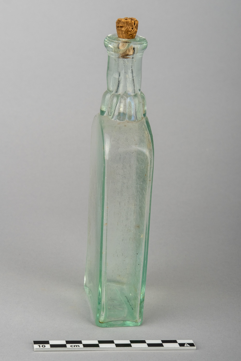 Flaske av blankt glass med svakt grønnaktig skjær. Rektangulær form med skrift på den ene bredsiden.