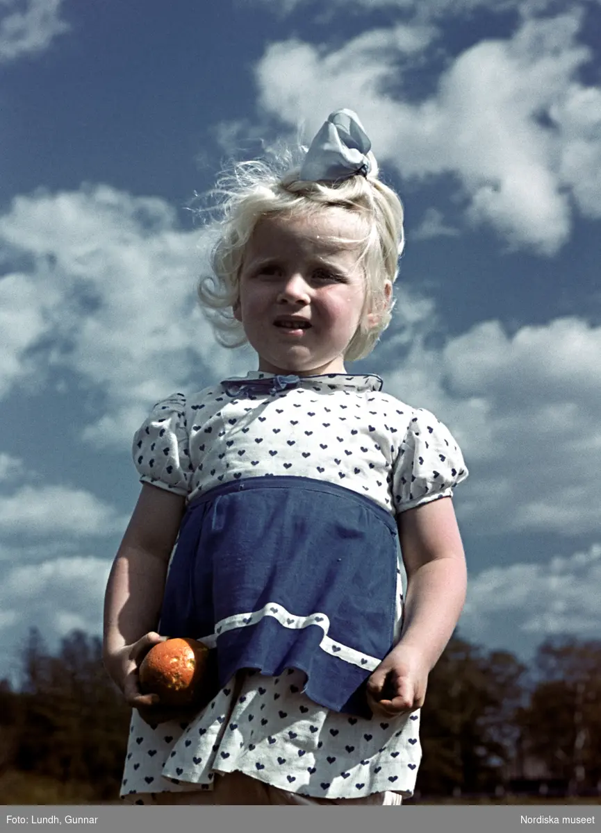 Flicka i vit klänning med hjärtmönster håller i ett äpple. Fotografed underifrån med himlen som bakgrund.