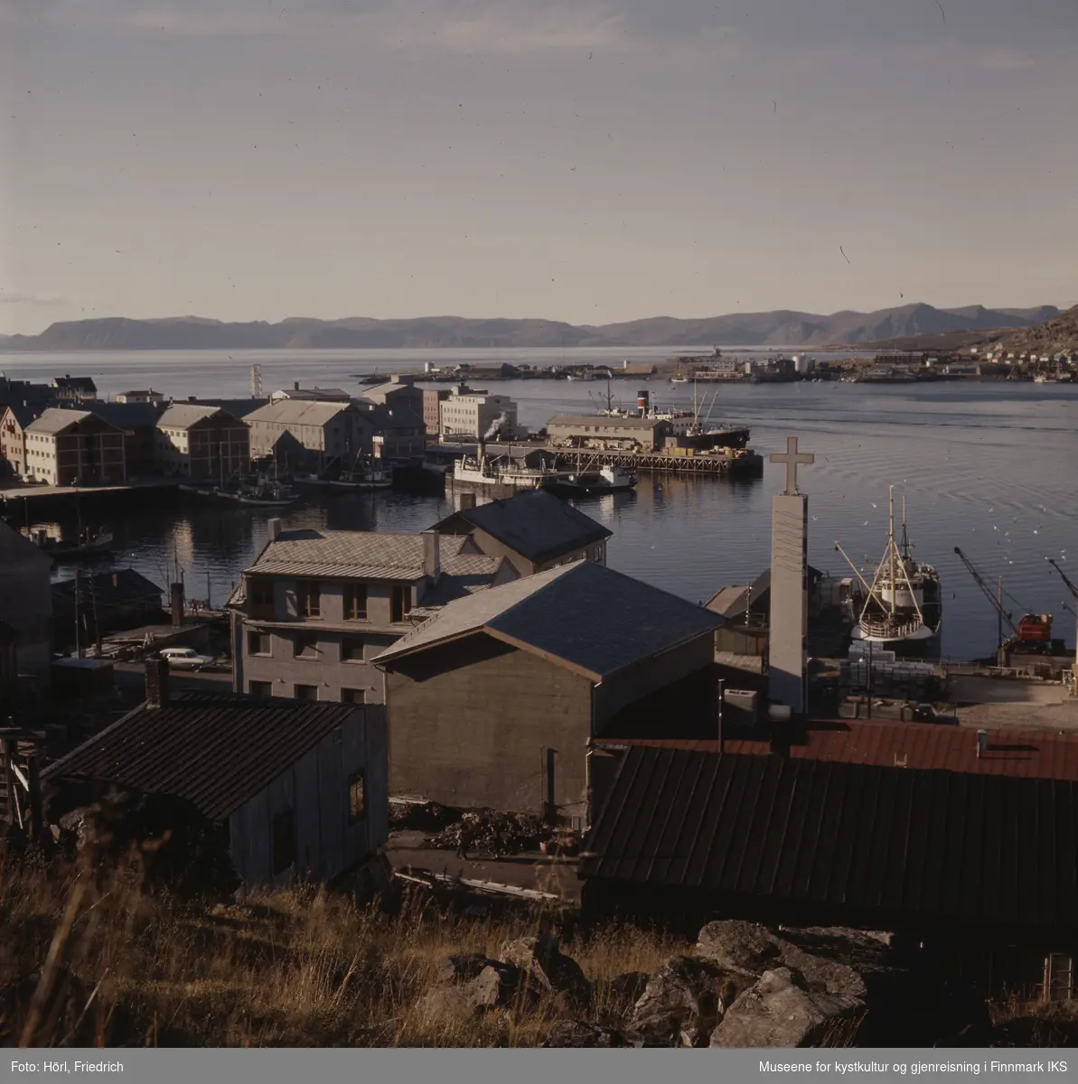 Utsikt over Hammerfest havn med deler av sentrumsbebyggelsen og båter i solskinn, fotografert fra Salen fjellet. I midten av bildet ser man den nybygde katolske kirka fra baksiden. Klokketårnet med sitt kors rager over takene. I bakgrunn ser man bydelen Fuglenes og Sørøysundet.