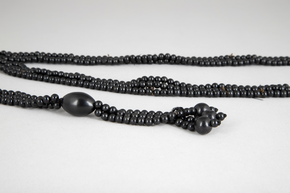 Langt, svart perlehalskjede med dusk nederst. Smykket er satt sammen av tre rader små perler av plastlignende materiale.