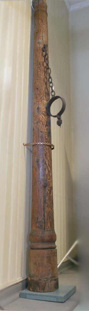 Brune trestokk med enkel utskåret profilering i form av runde bånd øverst og nedert. Påmontert halsjern med lenke og hengelås.