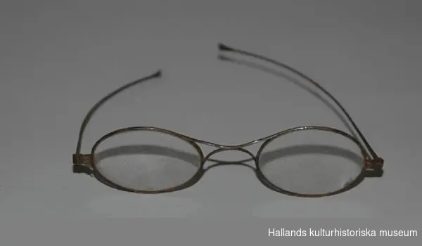 Glasögon i fordral av svart papper. Längd (fordral): 17 cm.Bredd (fordral): 3,5 cm.Längd (m. skalm): 10,8 cm.Längd (glaset): 3,8 cm.Bredd (glaset): 2,9 cm.