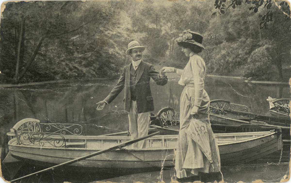 Postkort med motiv av ei kvinne og mann ved noen elvebåter.