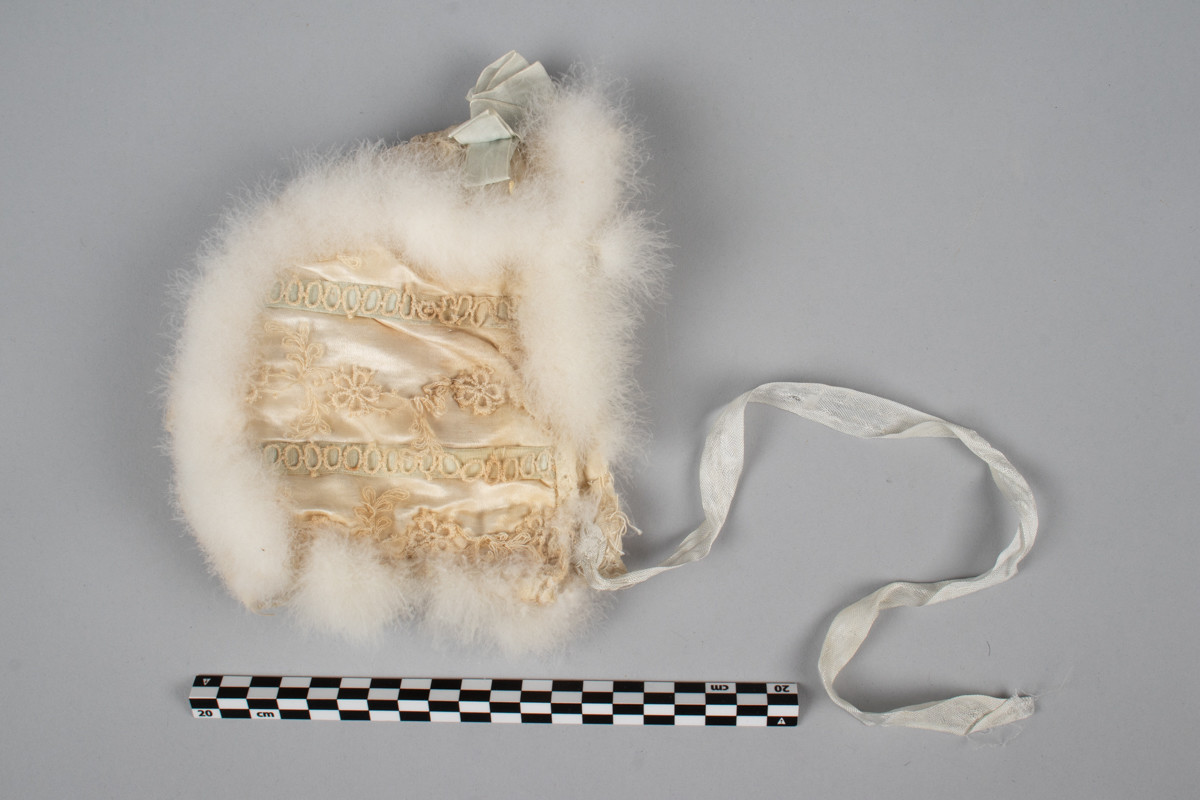 Liten dåpslue satt sammen av tre tøy stykker. Dekorert med påsydde nupereller, pels og lyseblå bånd