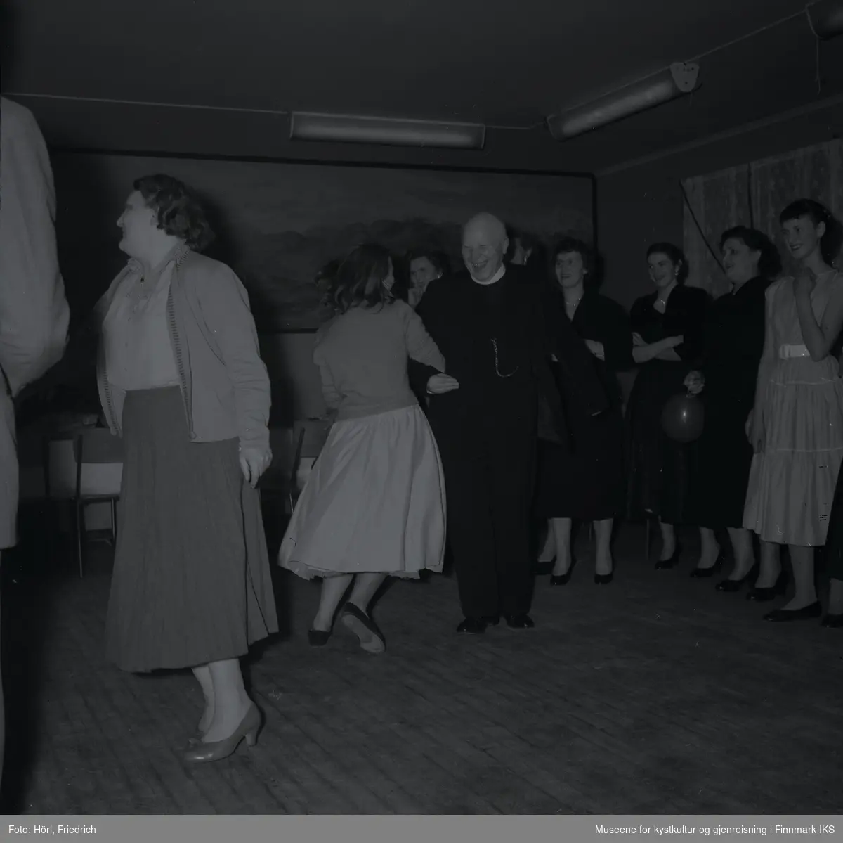 Den katolske menigheten i Hammerfest feirer juletrefest i 1957. Midt i bilde danser Pater Stockmann med en kvinne som har ryggen vendt til fotografen. På siden av dansegulvet er det flere kvinner som ser på sceneriet og er i godt humør.