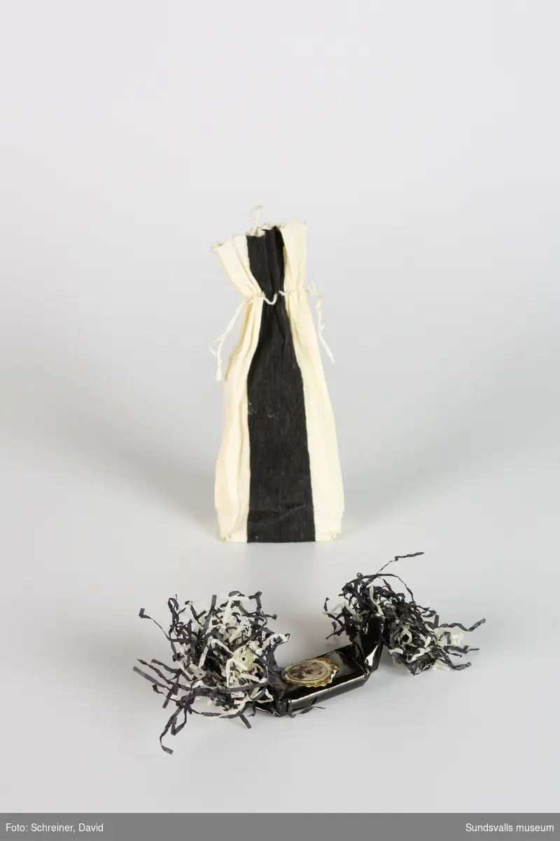 Påse tillverkad av svart- och vitrandigt crepepapper och dras ihop upptill med hjälp av ett vitt snöre. Påsen innehåller en begravningskaramell
