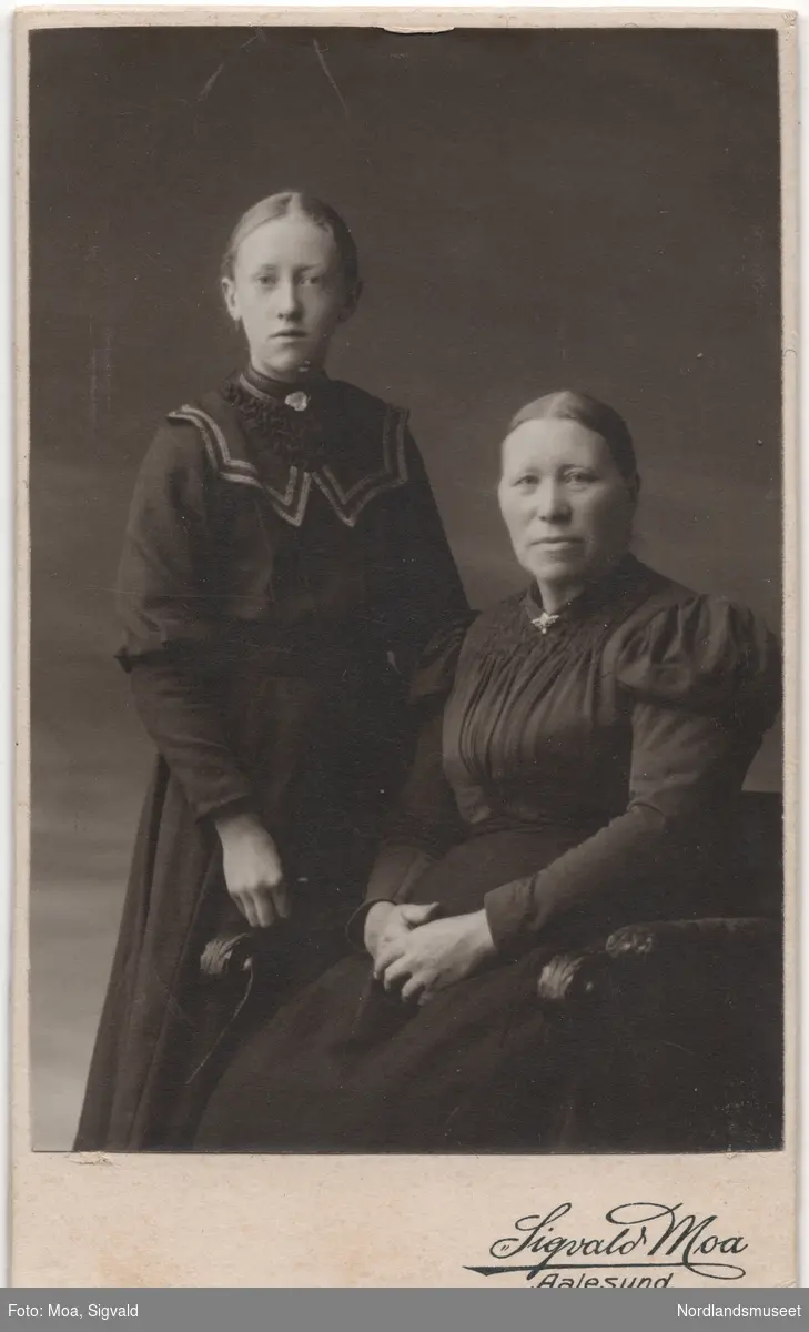 Portrett av en ung jente stående ved siden av en eldre kvinne sittende i en lenestol. Helga Fremmerlid med datter Marie, kalt Maja, fra Sunnmøre. Studiobilde. Helfigur.