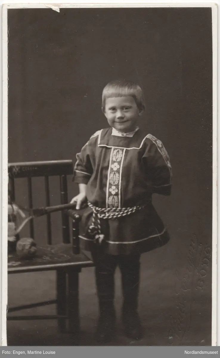 Portrett av et barn i en ornamentert kjole eller bluse, muligens litt inspirert av en samekofte elelr annen folkedrakt. Studiobilde. Gutten (?) står ved en stol. Helfigur.