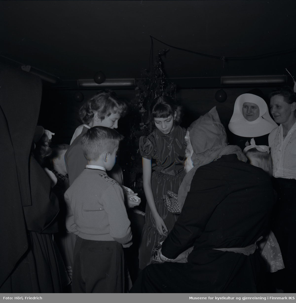 Julenissen er på besøk på juletrefesten til den katolske menigheten i Hammerfest i 1957. Barna mottar gaver mens de voksne, samt to nonner, ser på. Jenta til venstre i midten av bilde er Lillian Trondsen. Til høyre for henne står Kamilla Trondsen. I bakgrunnen ser man juletreet.