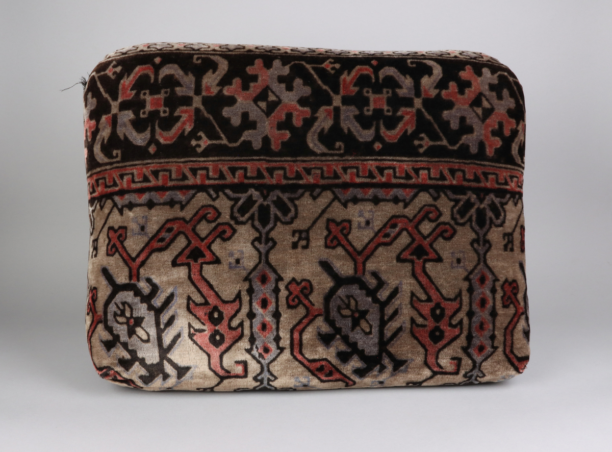 En kudde av mönstrad schagg. Orientaliserande mönster i färgerna grå, svart och vinröd. Del i en grupp med överkast och kuddar till en sk. Ottoman, en typ av bäddsoffa.
