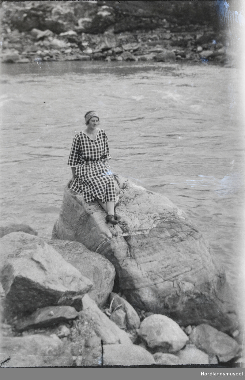 Kvinne i rutet kjole sitter på en stor stein ved bredden av en stri elv. Sannsynligvis Klara Bøe.